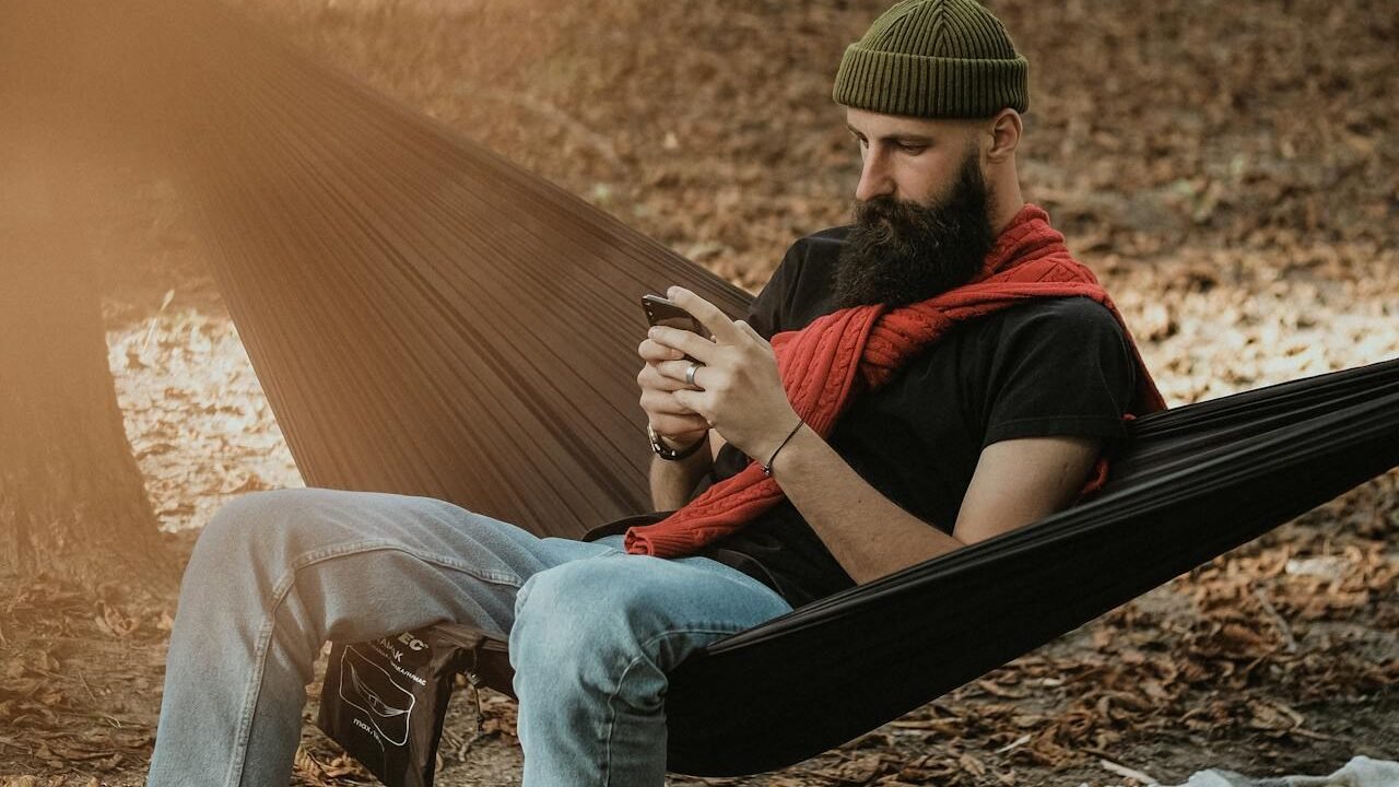 Hombre viendo el móvil sentado en una hamaca para colgar en el exterior.